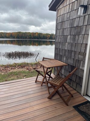 Una terraza de madera con una mesa y sillas, con vista a un lago tranquilo y follaje otoñal al fondo.