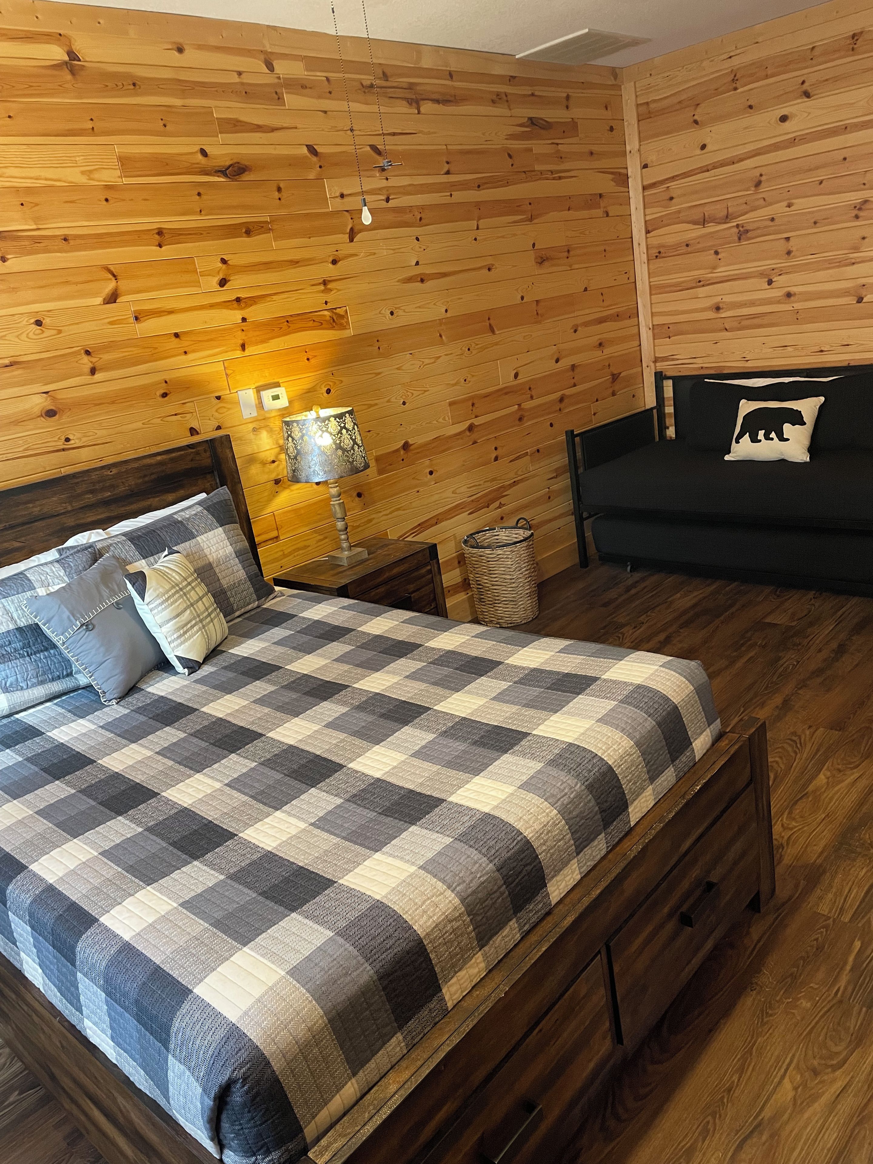 Ein Zimmer mit einer Holzwand, einem Bett mit kariertem Überwurf, einer Nachttischlampe, einer schwarzen Couch mit einem Bärenkissen und einem Korbwäscher.