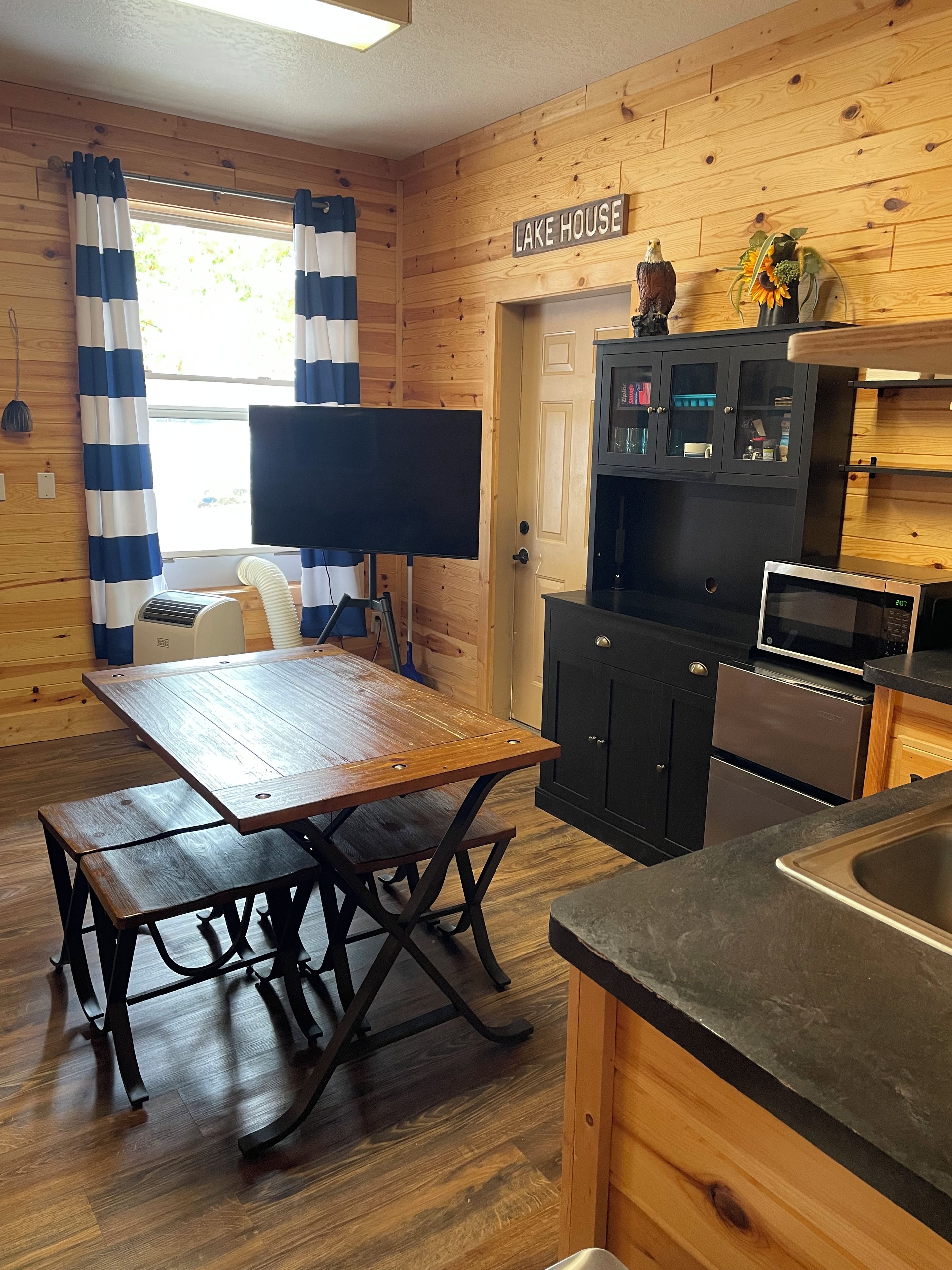 A wooden interior with a dining table, black cabinets, a microwave, and a sign reading 'LAKE HOUSE'.