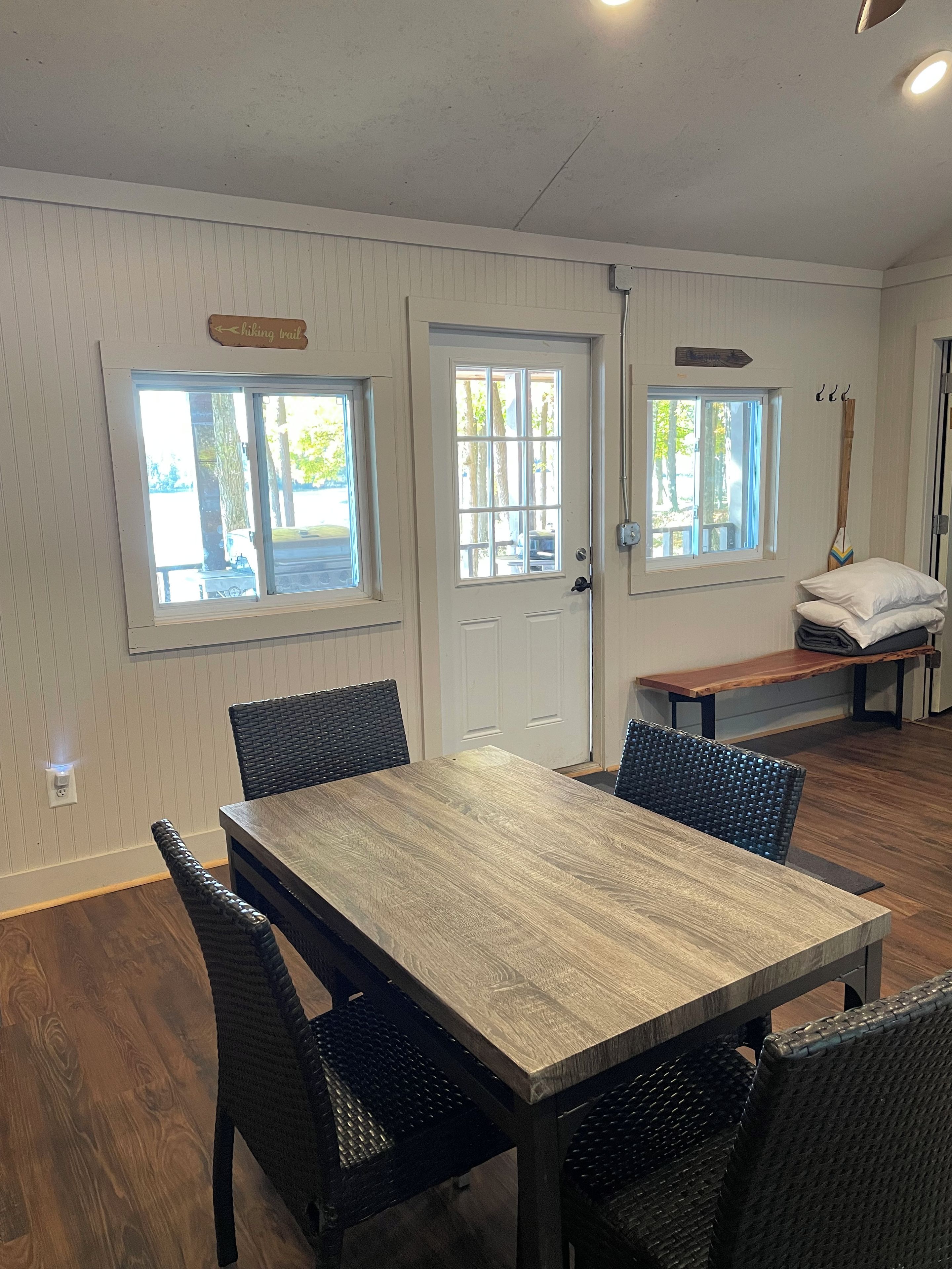Un comedor con mesa de madera y sillas de mimbre negro, un banco y dos ventanas con vistas a árboles.