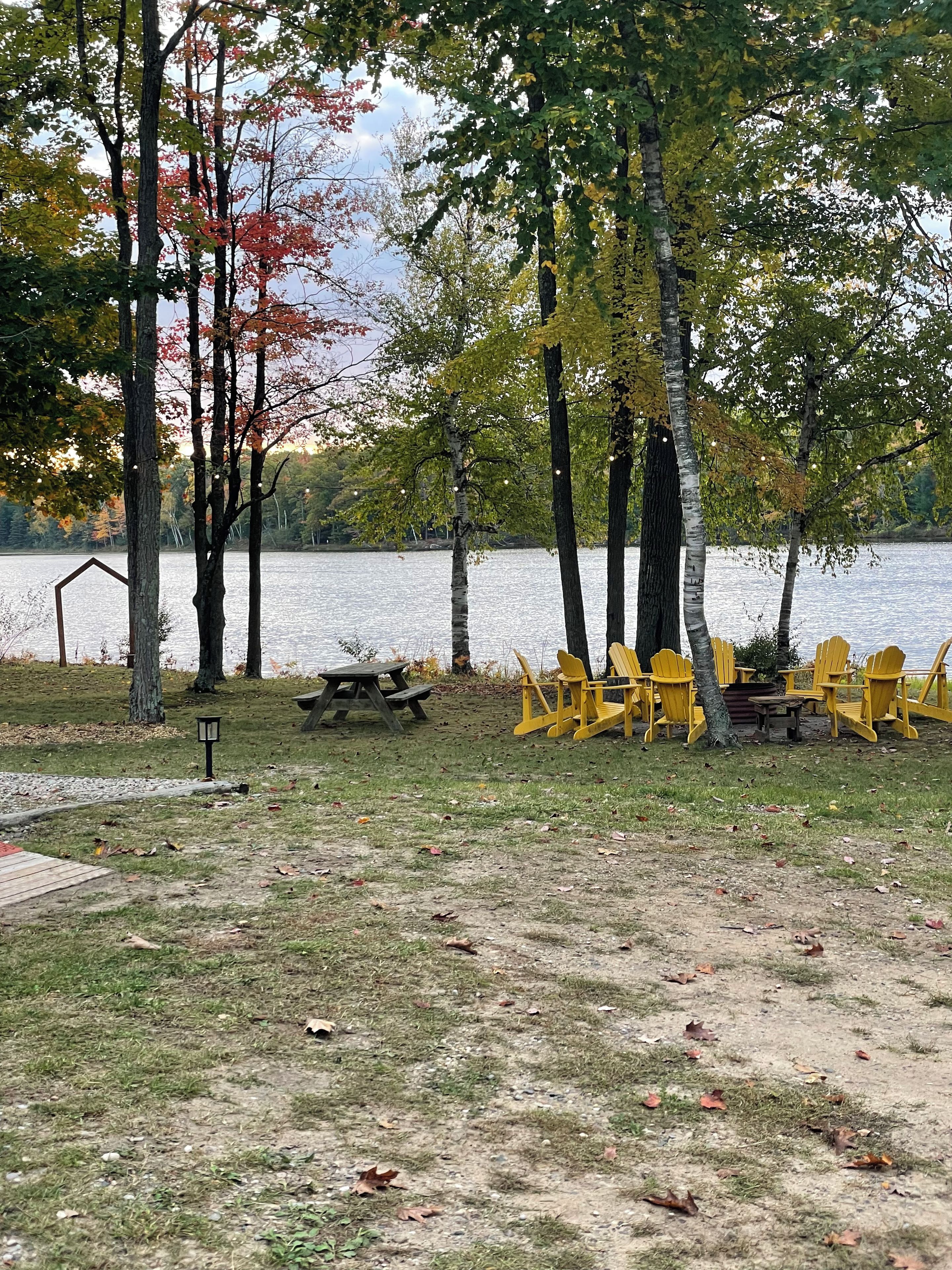 Un parque junto al lago con mesas de picnic, sillas Adirondack amarillas y árboles con follaje otoñal.
