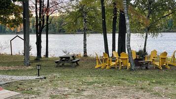 Un parque junto al lago con mesas de picnic, sillas Adirondack amarillas y árboles con follaje otoñal.