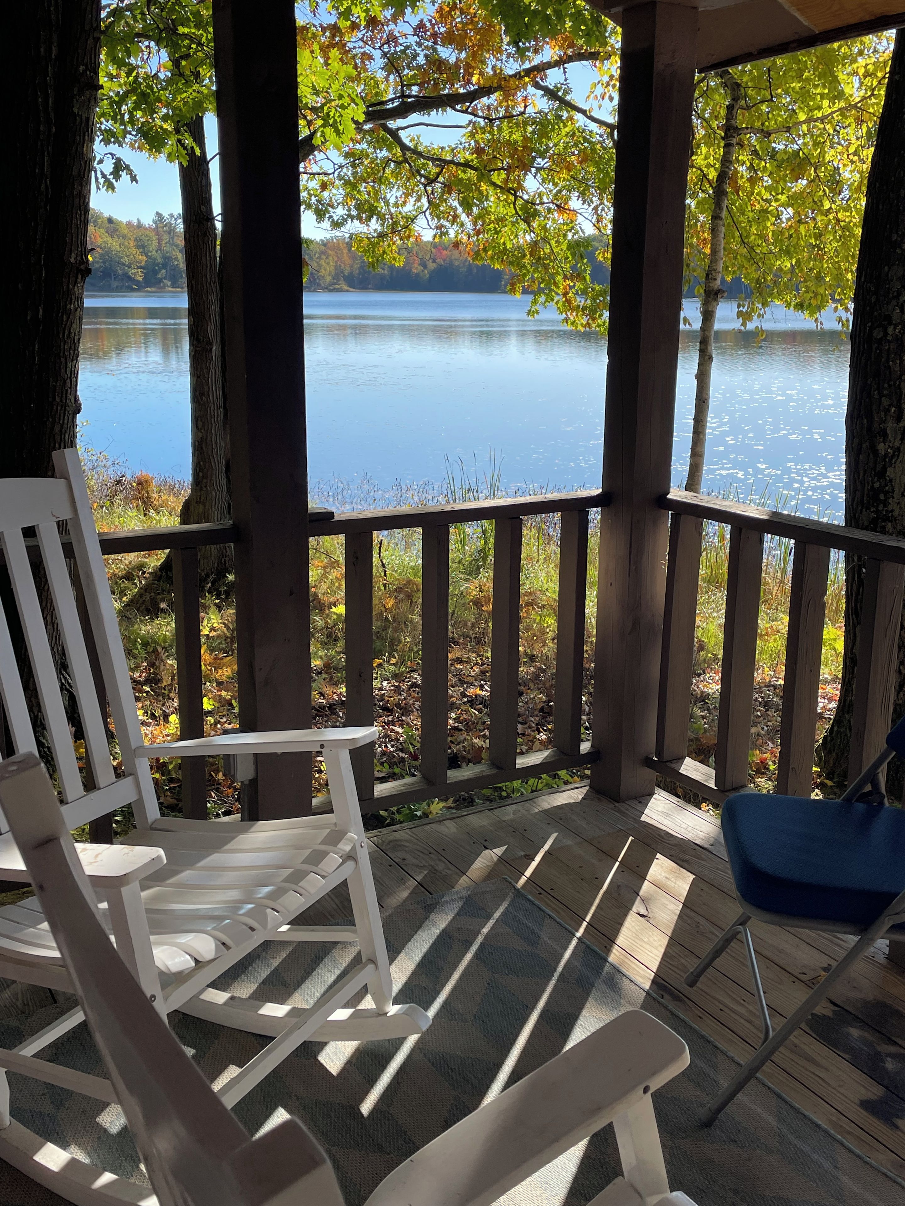 A wooden deck with a white rocking chair and a blue chair, overlooking a calm lake with trees in the background.