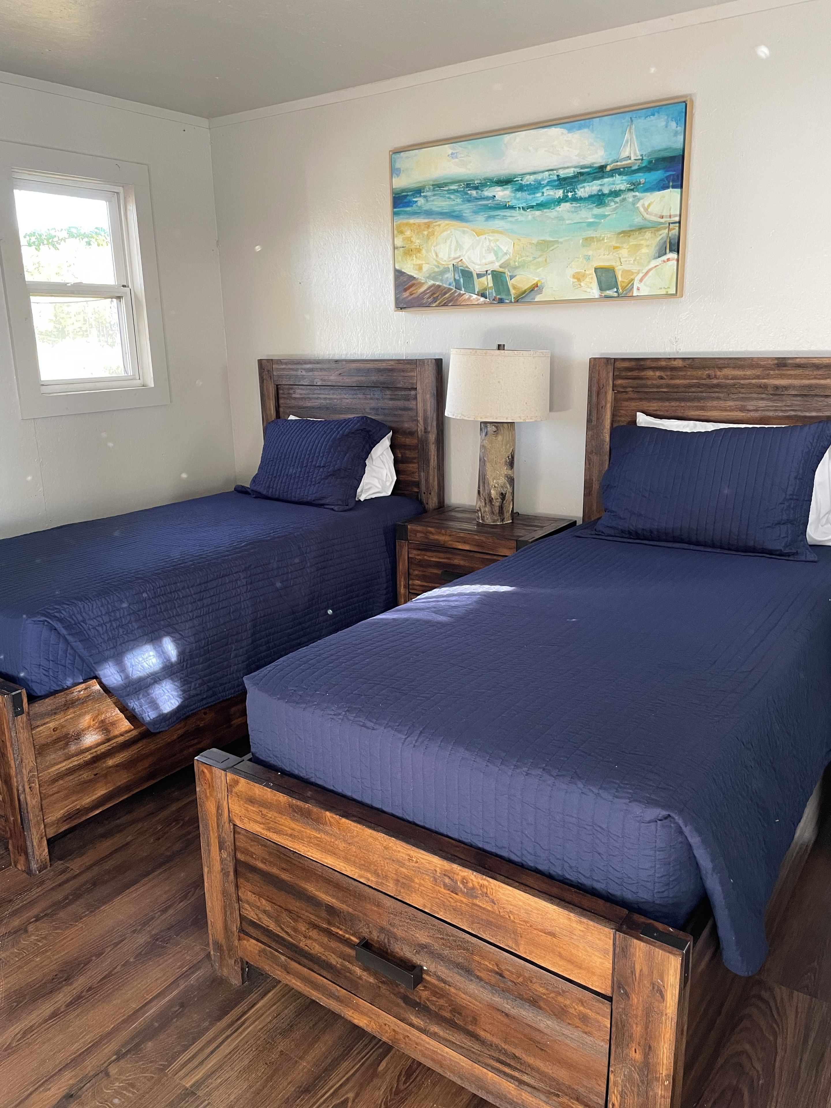 A bedroom with two wooden beds, a nightstand with a lamp, a painting on the wall, and a window.