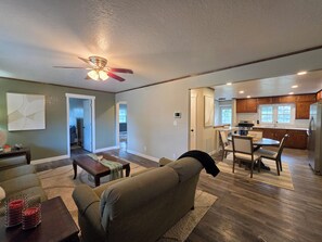 A spacious living room with a sofa, coffee table, and a ceiling fan. A kitchen with wooden cabinets and a dining area with a table and chairs is visible in the background.