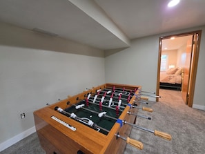 A foosball table in a room with a carpeted floor, a bed with a quilt, and a door leading to another room.