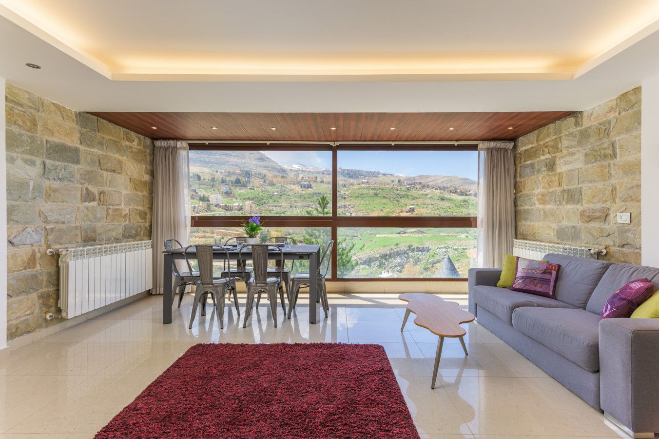 Amplia sala de estar con un ventanal que ofrece vistas panorámicas, un comedor con sillas, un sofá con cojines y una alfombra roja.