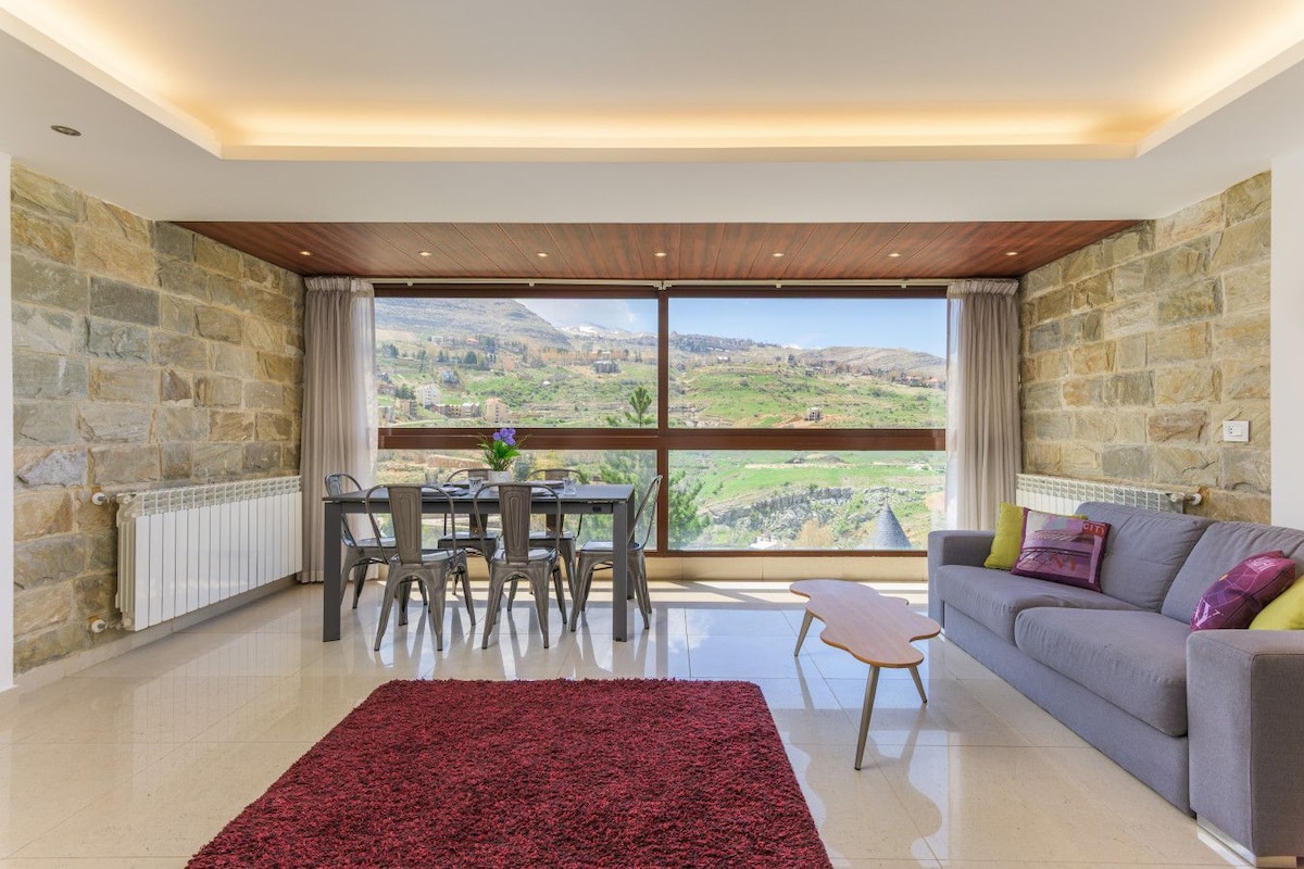 Ein geräumiges Wohnzimmer mit großem Fenster und herrlicher Aussicht, einem Essbereich mit Stühlen, einer Couch mit Kissen und einem roten Teppich.