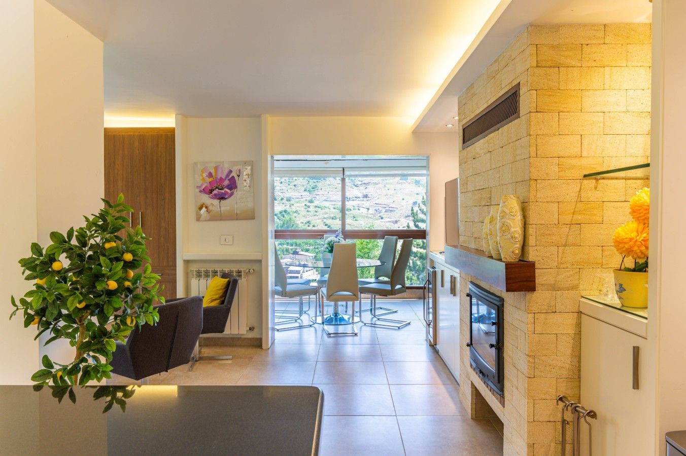 A modern living room with a fireplace, a dining area with a view, and a kitchen with a stone wall.