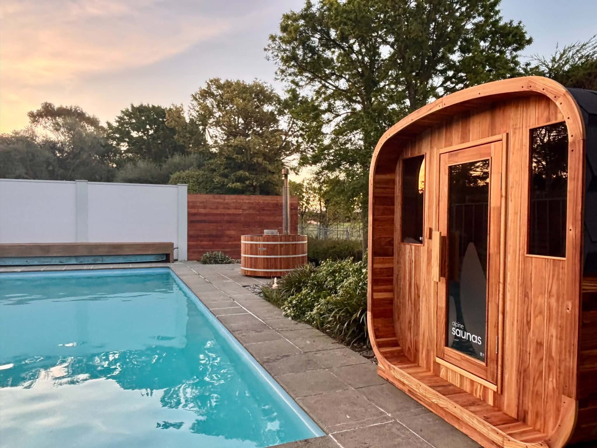 Estructura de sauna de madera junto a una piscina, con cielo despejado al fondo.