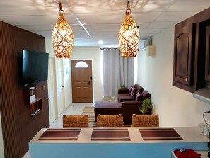 Ein modernes Wohnzimmer mit Flachbildfernseher, braunem Sofa, Korbstühlen und zwei Pendelleuchten.