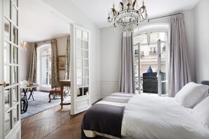 Een hotelkamer met een groot bed, een kroonluchter, een balkon met gordijnen en uitzicht op de stad.