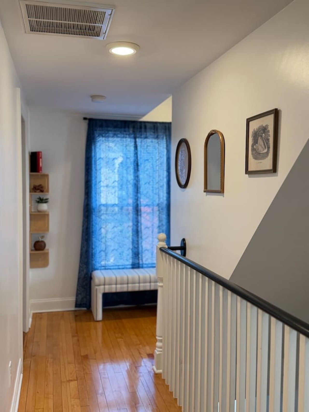 Un couloir doté d’un plancher en bois, d’un banc, d’un escalier avec une rampe et d’une fenêtre ornée de rideaux bleus.