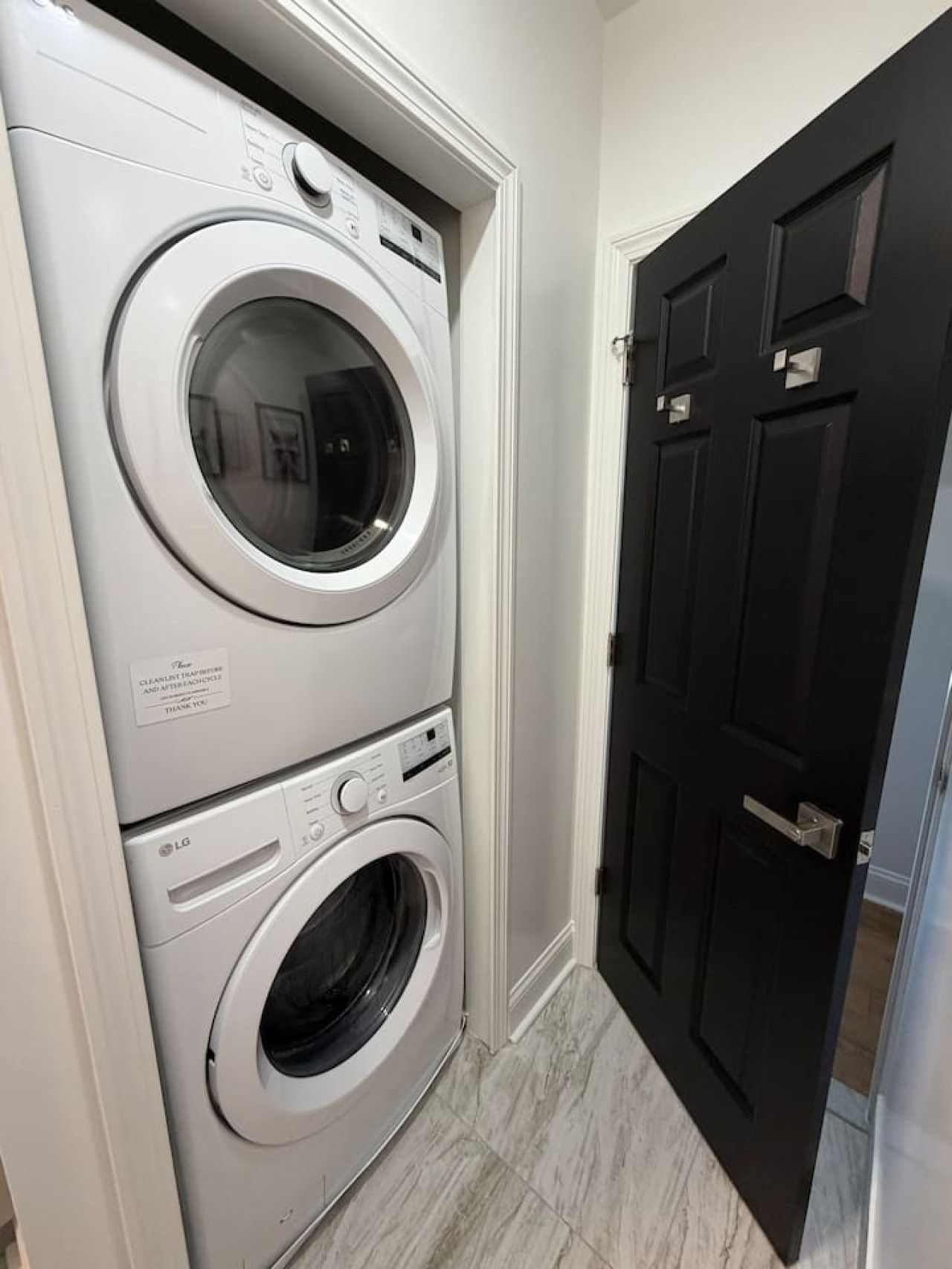 Une machine à laver et un sèche-linge superposés, installés dans un couloir avec une porte noire.