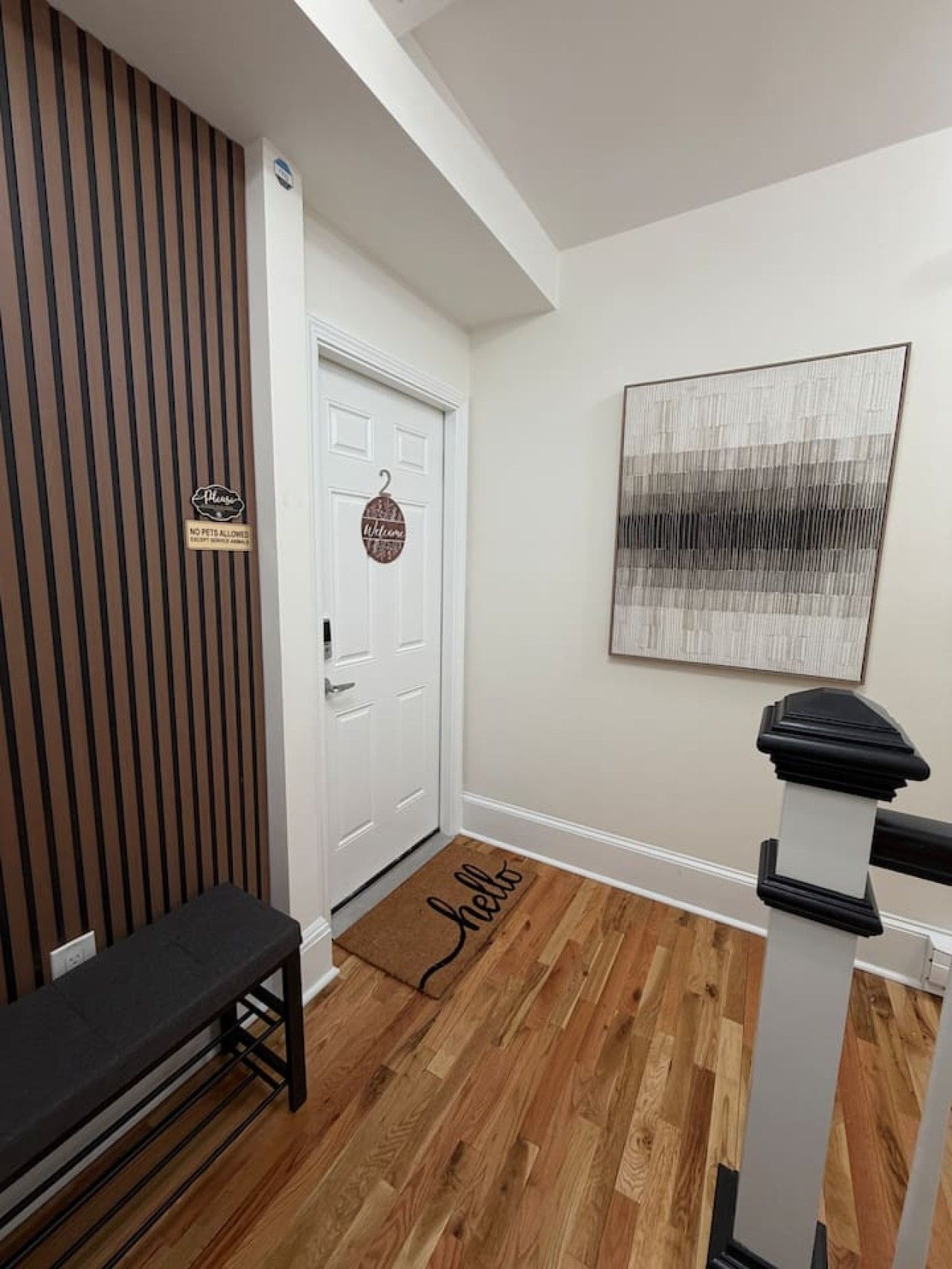A hallway with a wooden bench, a white door with a 'Welcome' sign, a doormat with 'hello' written on it, and a modern abstract painting on the wall.