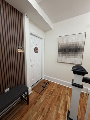 Ein Flur mit einer hölzernen Bank, einer weißen Tür mit einem 'Welcome'-Schild, einer Fußmatte mit der Aufschrift 'hello' und einem modernen, abstrakten Gemälde an der Wand.