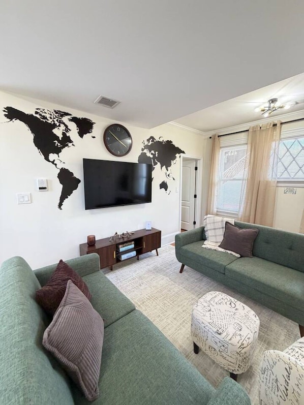 Una sala de estar con un televisor de pantalla plana grande, un mapa del mundo como decoración de pared, un sofá, una mesa de centro y una ventana con cortinas.