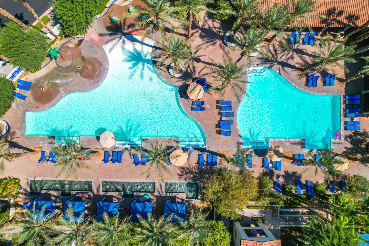 Vista aérea de un complejo turístico con dos piscinas grandes, rodeado de palmeras y sillas de descanso.