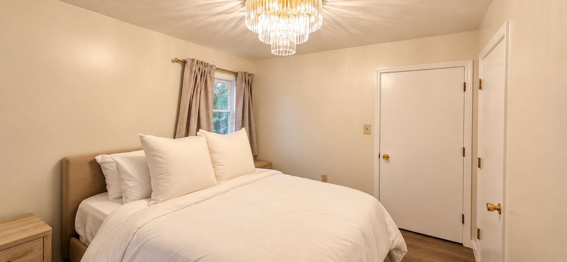 Ein Schlafzimmer mit einem Bett, einem Nachttisch, einem Fenster mit Vorhängen, einer Tür und einer Chandelier.