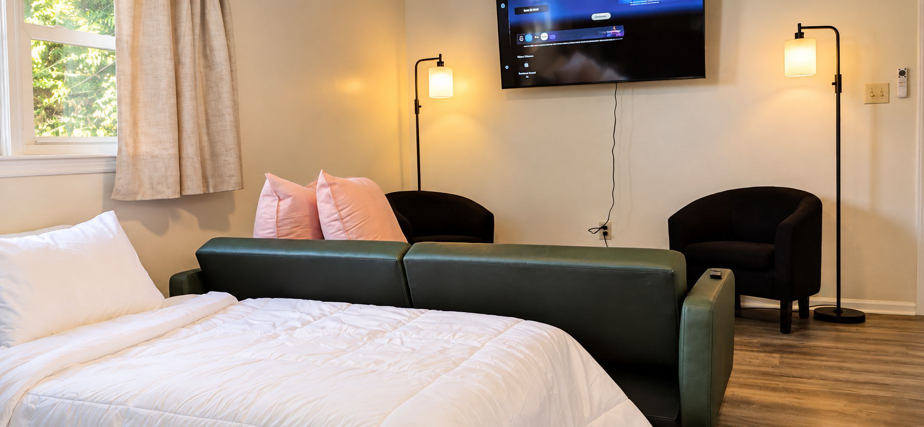 Ett hotellrum med en säng, två stolar, en tv som är monterad på väggen och ett fönster med gardiner.