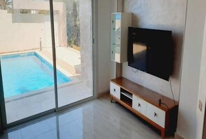 Une chambre moderne dotée d’une grande porte-fenêtre coulissante donnant sur une piscine, d’un téléviseur à écran plat fixé au mur et d’un meuble en bois avec des tiroirs.