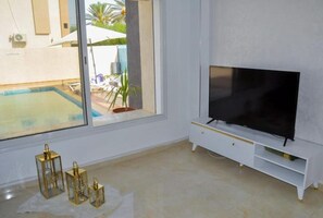 Une pièce dotée d’une grande fenêtre, d’un téléviseur à écran plat posé sur un meuble blanc, et d’où l’on peut voir une piscine à l’extérieur.