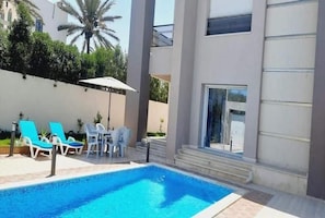 Ein Pool mit Liegestühlen und einem Tisch, umgeben von einem Gebäude mit großen Fenstern und einer Palme im Hintergrund.