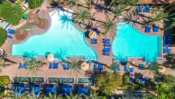 An aerial view of a resort with two large swimming pools, surrounded by palm trees and lounge chairs.
