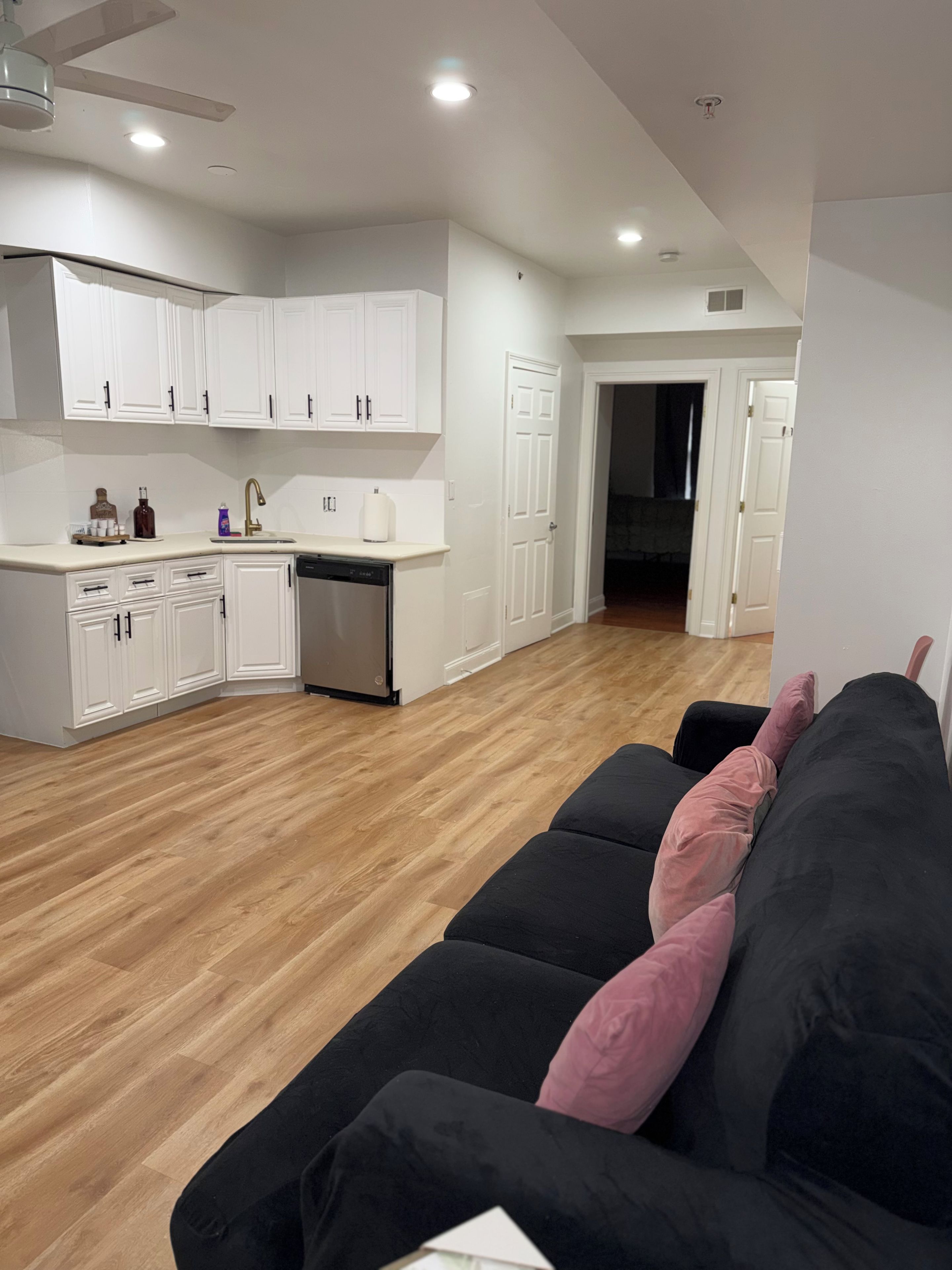 Una cocina moderna con armarios blancos y lavavajillas, un sofá negro con almohadas rosas y piso de madera.