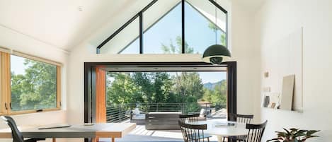 A modern interior with a dining area, a desk, and a large window offering a view of greenery and mountains.