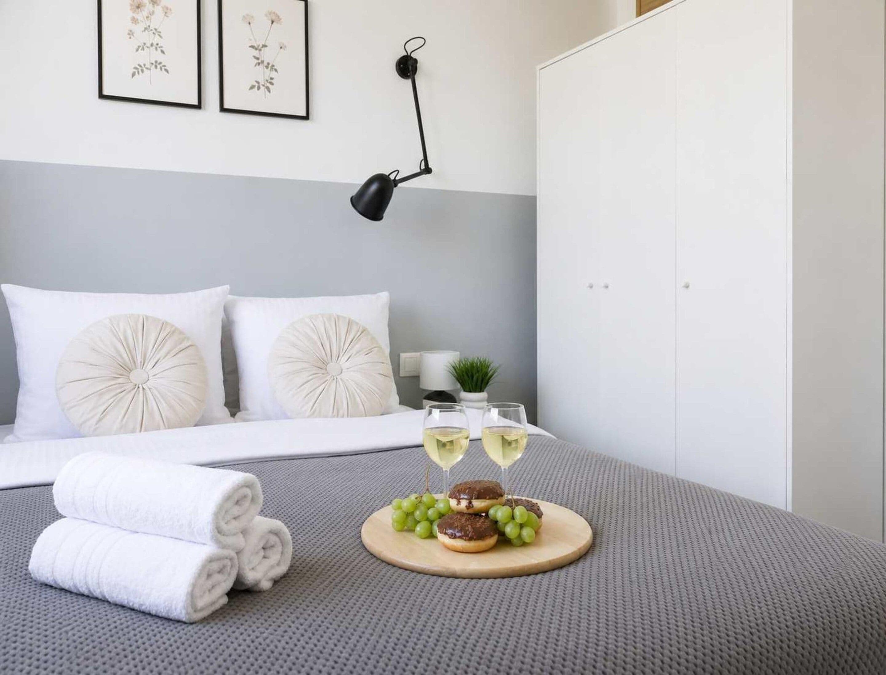 Een net opgemaakt bed met witte lakens, een paar opgerolde handdoeken, een houten dienblad met snacks en glazen witte wijn, en een wandlamp.