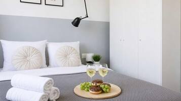 Een net opgemaakt bed met witte lakens, een paar opgerolde handdoeken, een houten dienblad met snacks en glazen witte wijn, en een wandlamp.