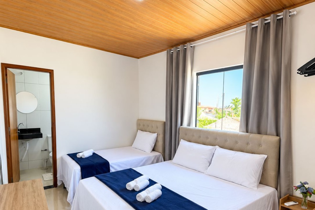 A hotel room with two beds, a wooden ceiling, a view of the outdoors, and a small bedside table with a vase.