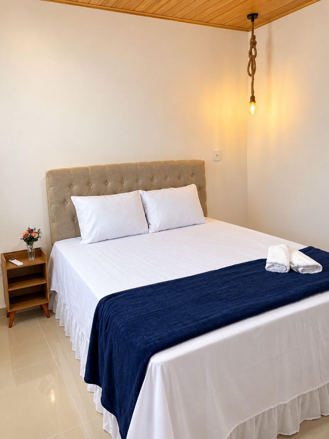 A neatly made bed with white linens and a dark blue throw blanket, a bedside table with a vase of flowers, and a wall-mounted light fixture.