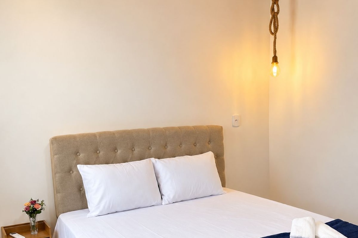 A neatly made bed with white linens and a dark blue throw blanket, a bedside table with a vase of flowers, and a wall-mounted light fixture.