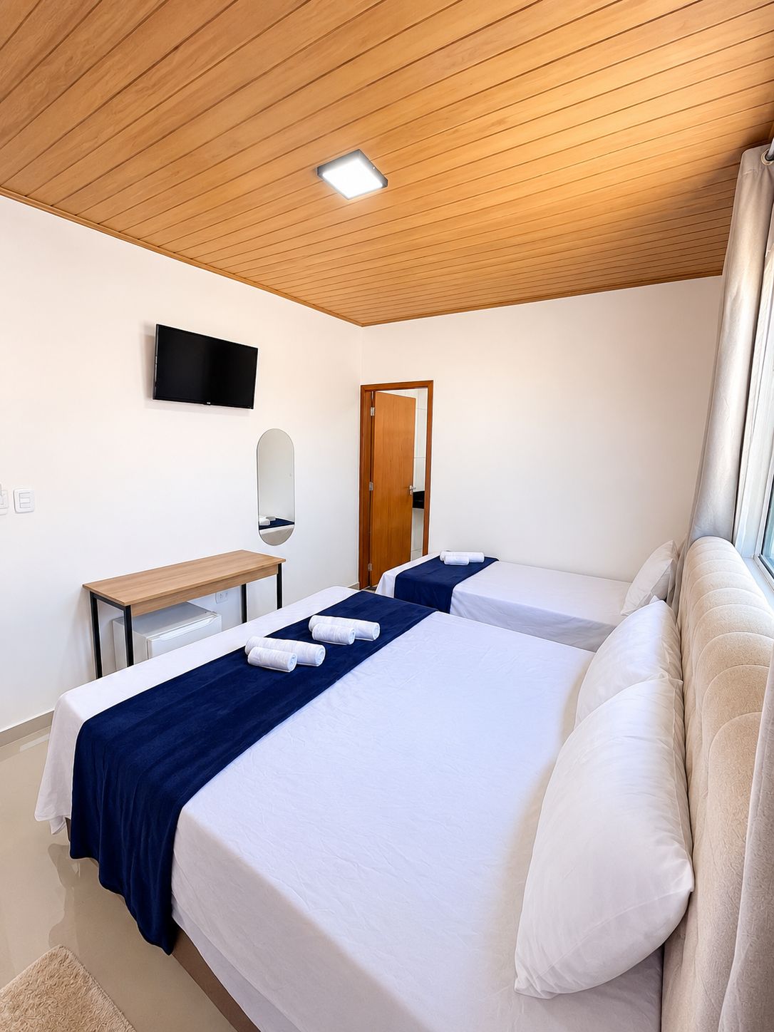 Habitació d'hotel amb dos llits, un televisor de pantalla plana muntat a la paret, sostre de fusta i una porta que condueix a una altra habitació.