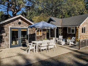 A wooden cabin with a patio area featuring a table and chairs, a covered seating area, and a small deck. - Two Stylish Cottages, One Coastal Escape | Walk to Beach | Dog Friendly (Cannon Beach)
