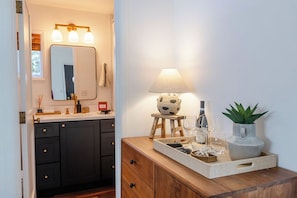 A bathroom with a dark wooden vanity, a mirror, a light fixture, and a small stool with a bowl and a plant. - Two Stylish Cottages, One Coastal Escape | Walk to Beach | Dog Friendly (Cannon Beach)