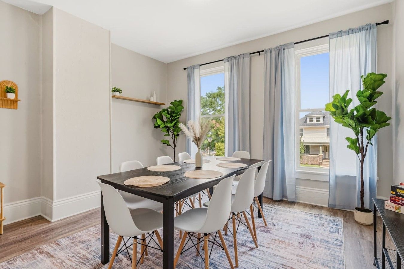 Un comedor con una mesa de madera oscura, sillas blancas y vistas al exterior a través de ventanas amplias con cortinas azul claro.