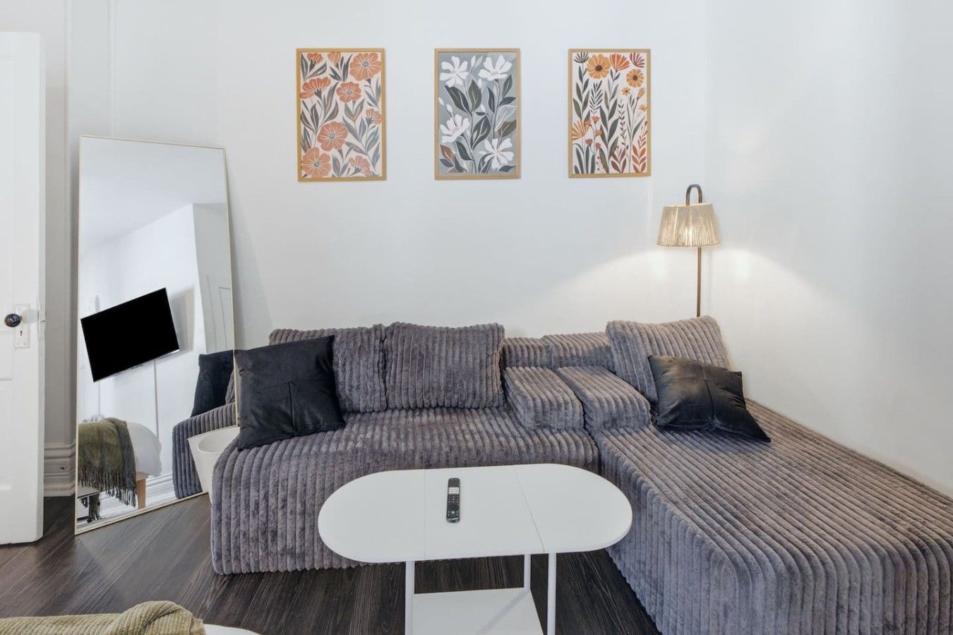 Un saló modern amb un sofà gris, una taula petita blanca i una làmpada de peu. Hi ha tres quadres florals amb marc a la paret.