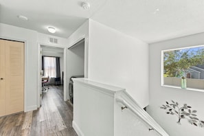 Un couloir doté d’un plancher en bois, d’un mur blanc, d’une fenêtre donnant sur un espace verdoyant et d’une porte à panneaux ajourés.