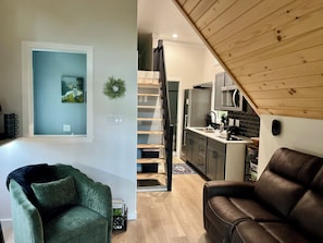 A modern living room with a green armchair, a brown leather sofa, a staircase leading to a kitchen with a microwave, and a small dining area.