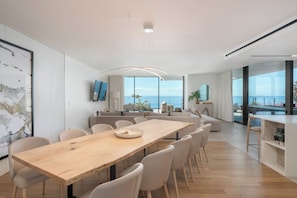 A modern dining area with a wooden table and chairs, a large window with a view of the sea, and a living area with a sofa and a television. - Savoy Luxe - Luxury 3-bed w/ pool (Funchal)