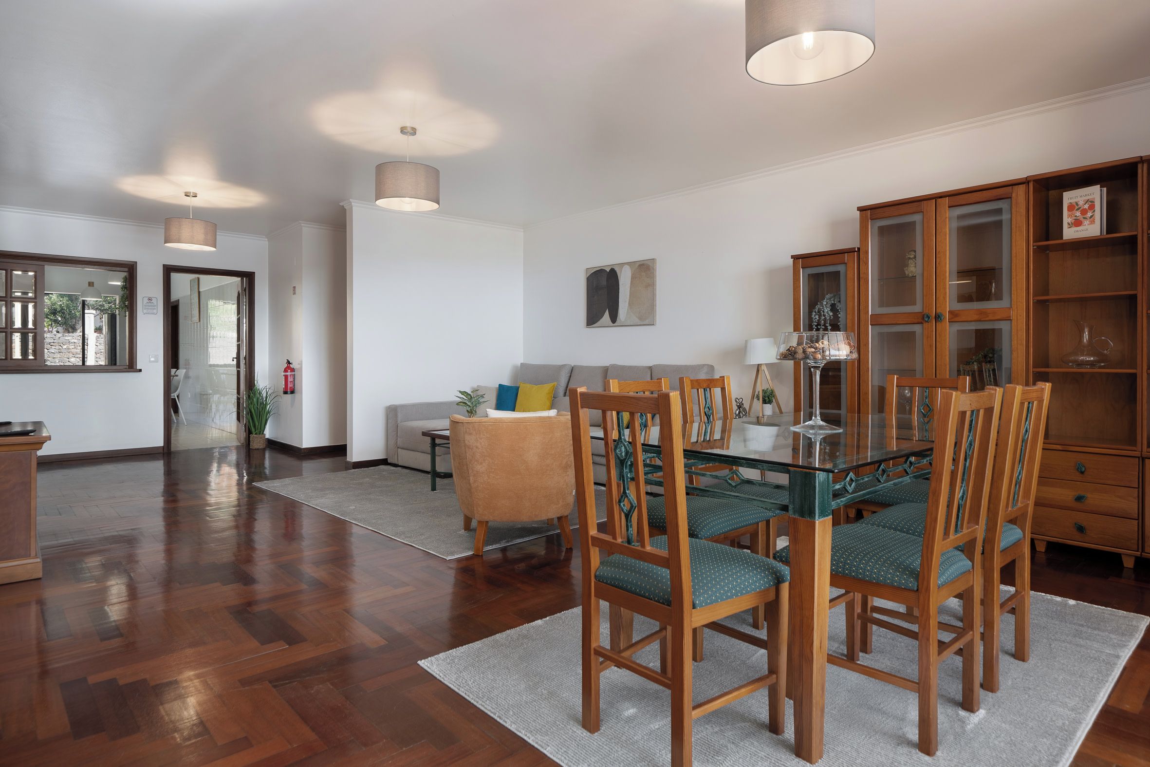 Un coin repas avec une table en verre et des chaises en bois, un buffet en bois doté de portes en verre, et un canapé à l’arrière-plan.