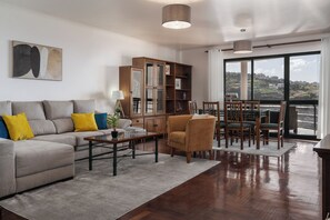 A living room with a sofa, coffee table, and chairs, a bookshelf, and a view through a large window.