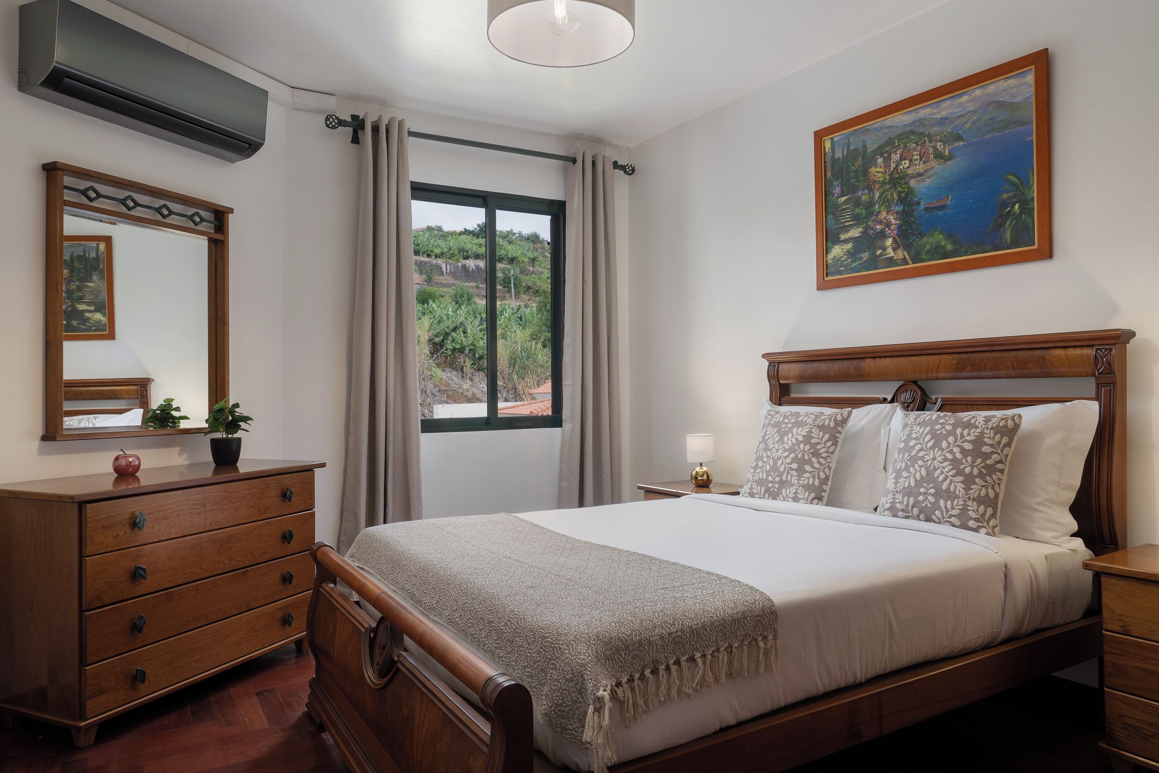 A bedroom with a wooden bed, a nightstand, a dresser, a mirror, a window with curtains, and a painting on the wall.