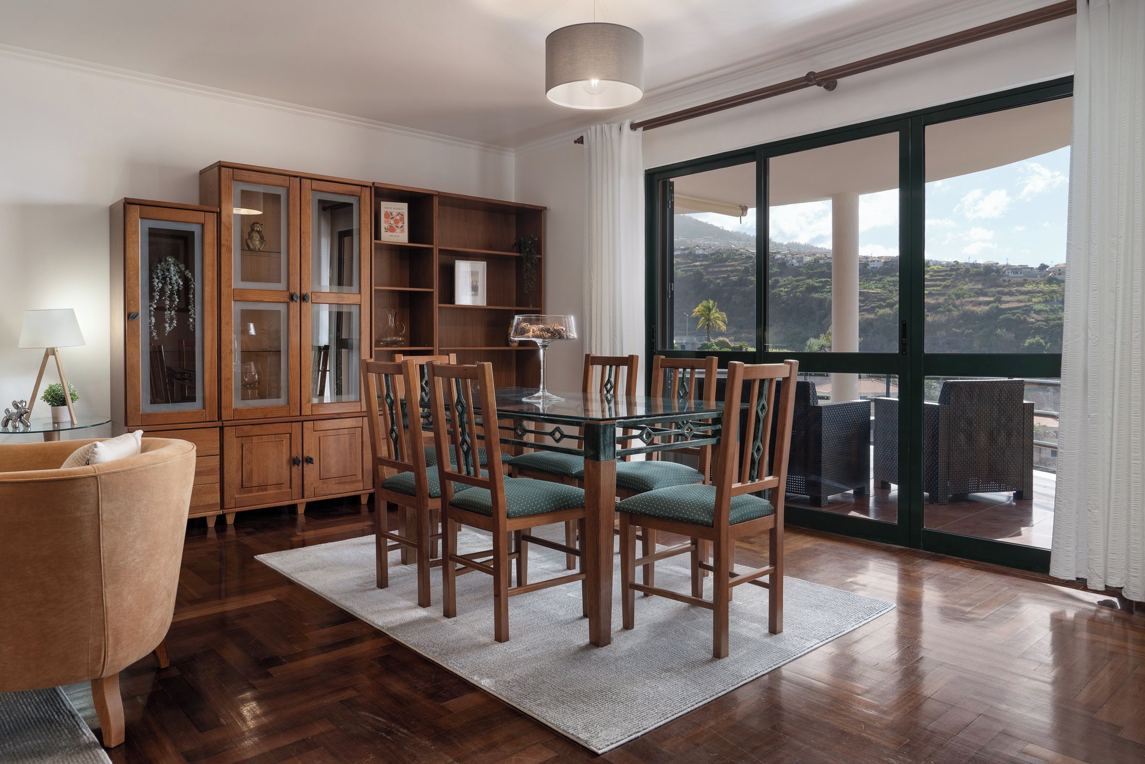 Un comedor con mesa de vidrio y sillas de madera, un mueble de madera con puertas de vidrio, y vista a través de un ventanal amplio.