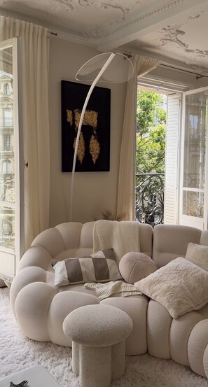 Een gezellige woonkamer met een ruime, beige hoekbank, een kunstwerk in zwart en goud, en een wit plafond met decoratieve ornamenten.