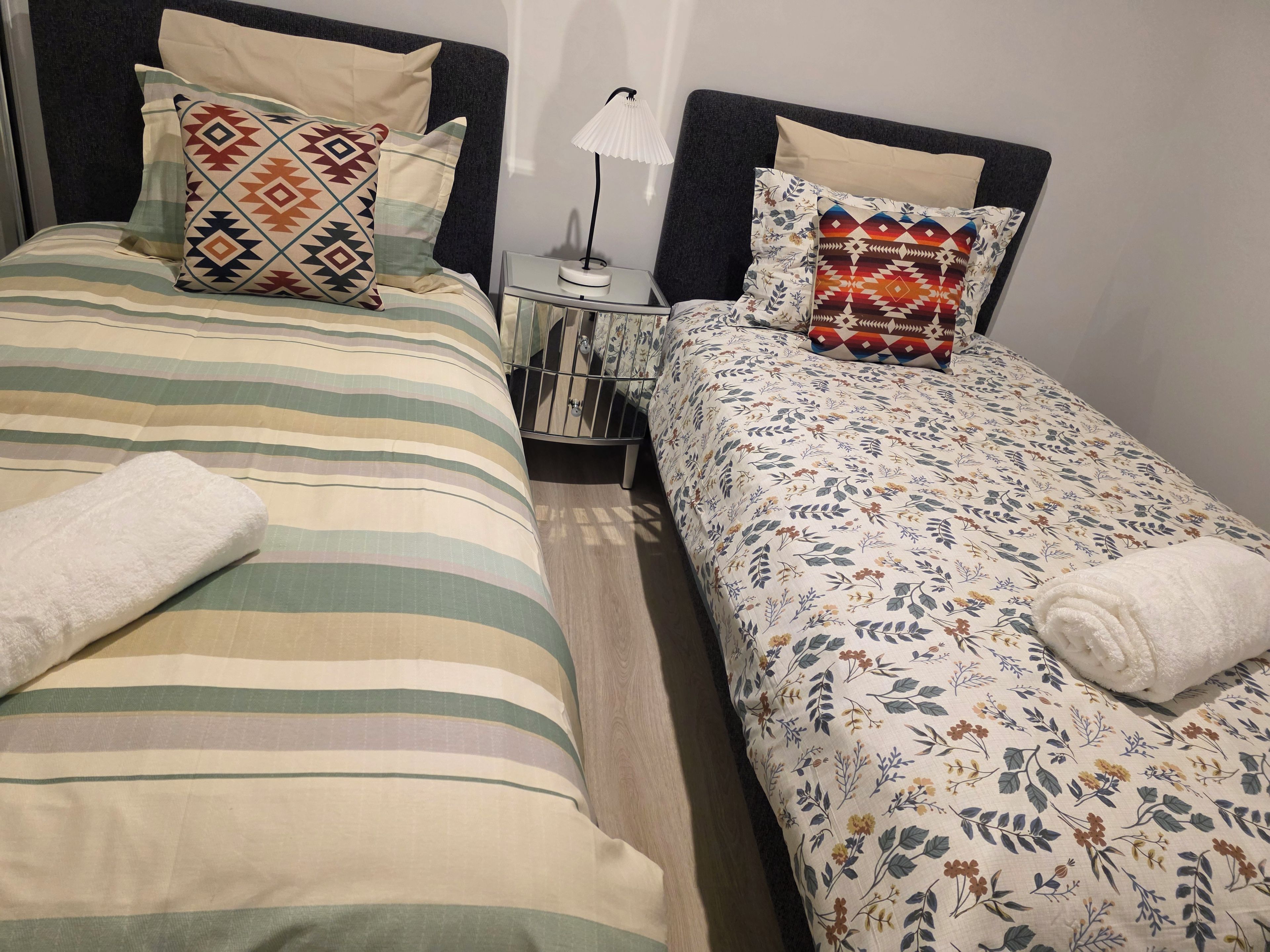 A hotel room with two beds, each with a different patterned bedspread, a bedside table with a lamp, and a mirror.
