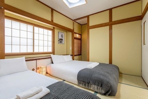 Ein traditioneller japanischer Raum mit Tatami-Bodenbelag, zwei Betten mit weißer Bettwäsche, einem Shoji-Bildschirm und einem kleinen Tisch mit einer Lampe.